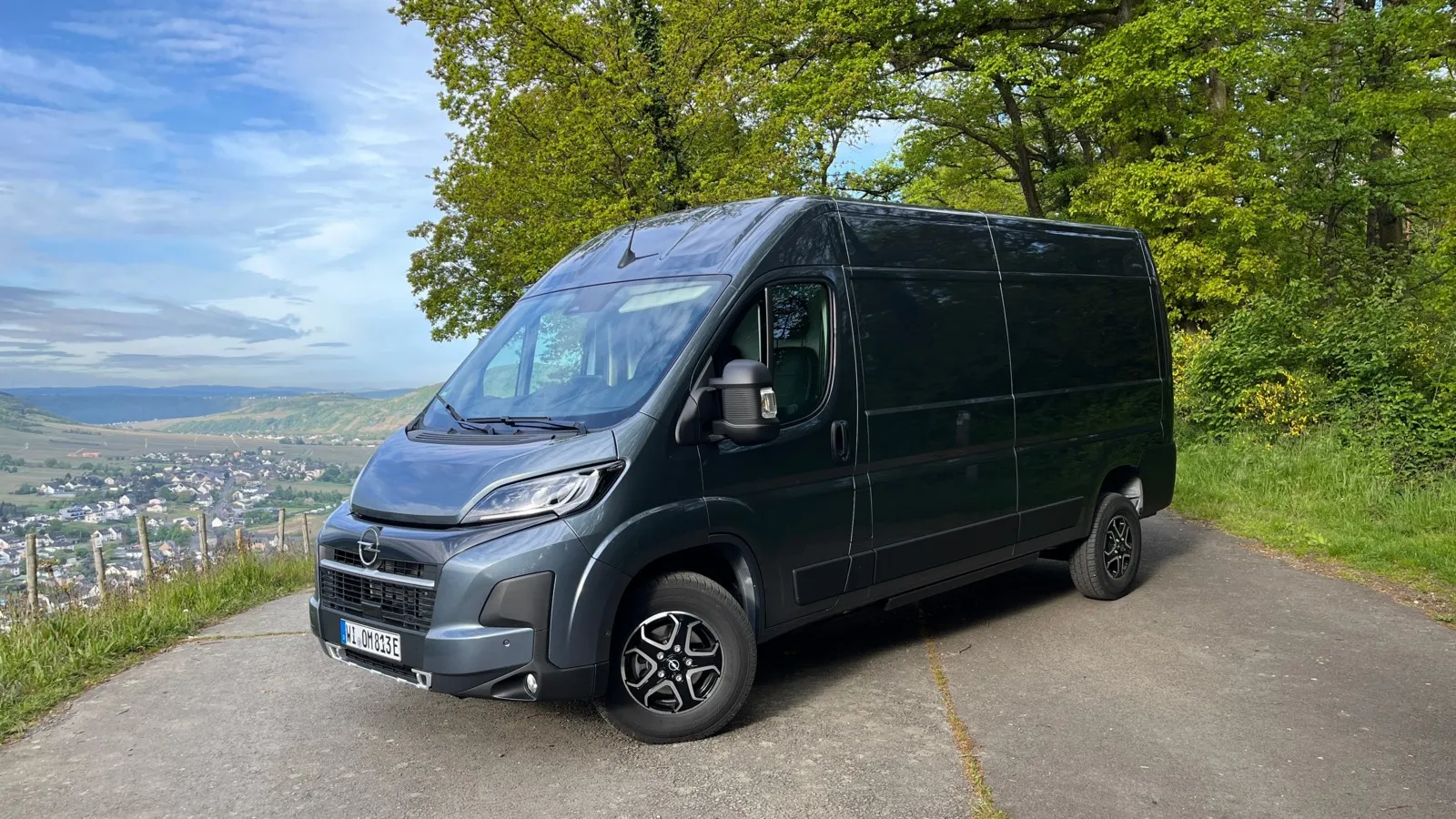 Frontansicht: Der Opel Movano Electric in dunkelgrau steht vor einem Baum.